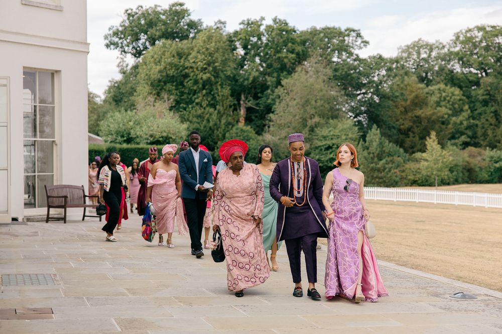 Wedding guests in colorful pink and purple traditional attire walk along a terrace at an outdoor venue.