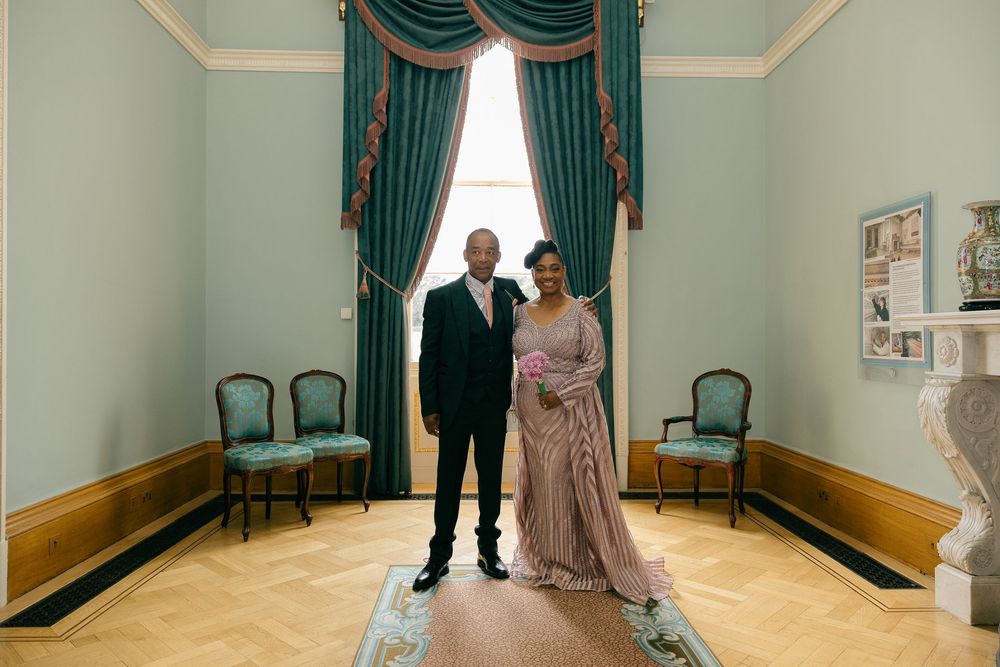 Couple in formal attire posing in elegant room with teal curtains and parquet floors.