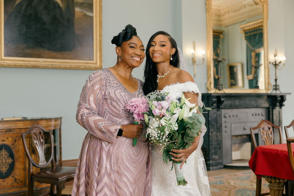 Bride in white off-shoulder gown holds bouquet beside woman in pink beaded dress, posing in elegant blue room.