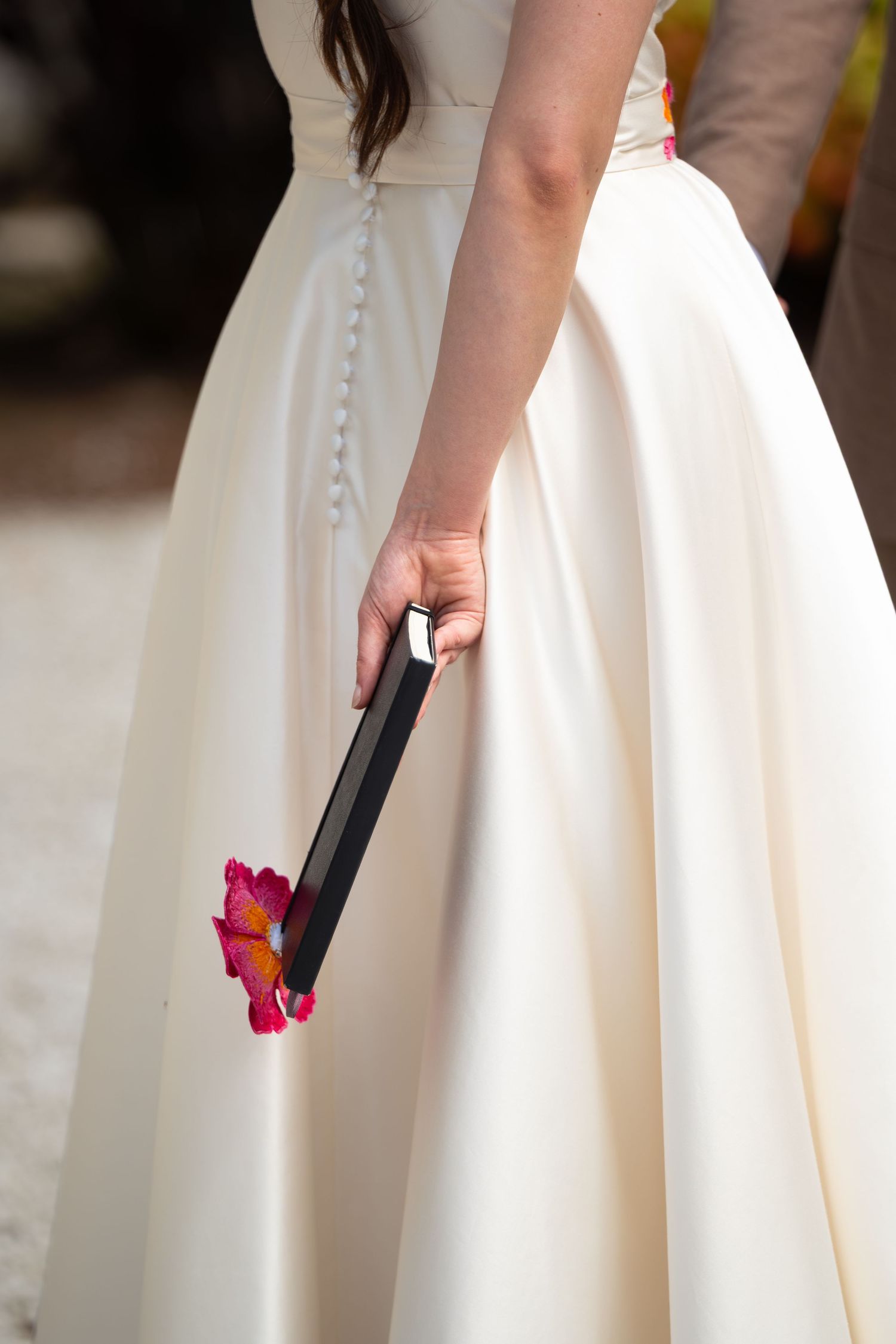 La mariée en robe écrue en satin tenant un carnet noir avec une fleur rose orangée brodée assortie à la ceinture de sa robe