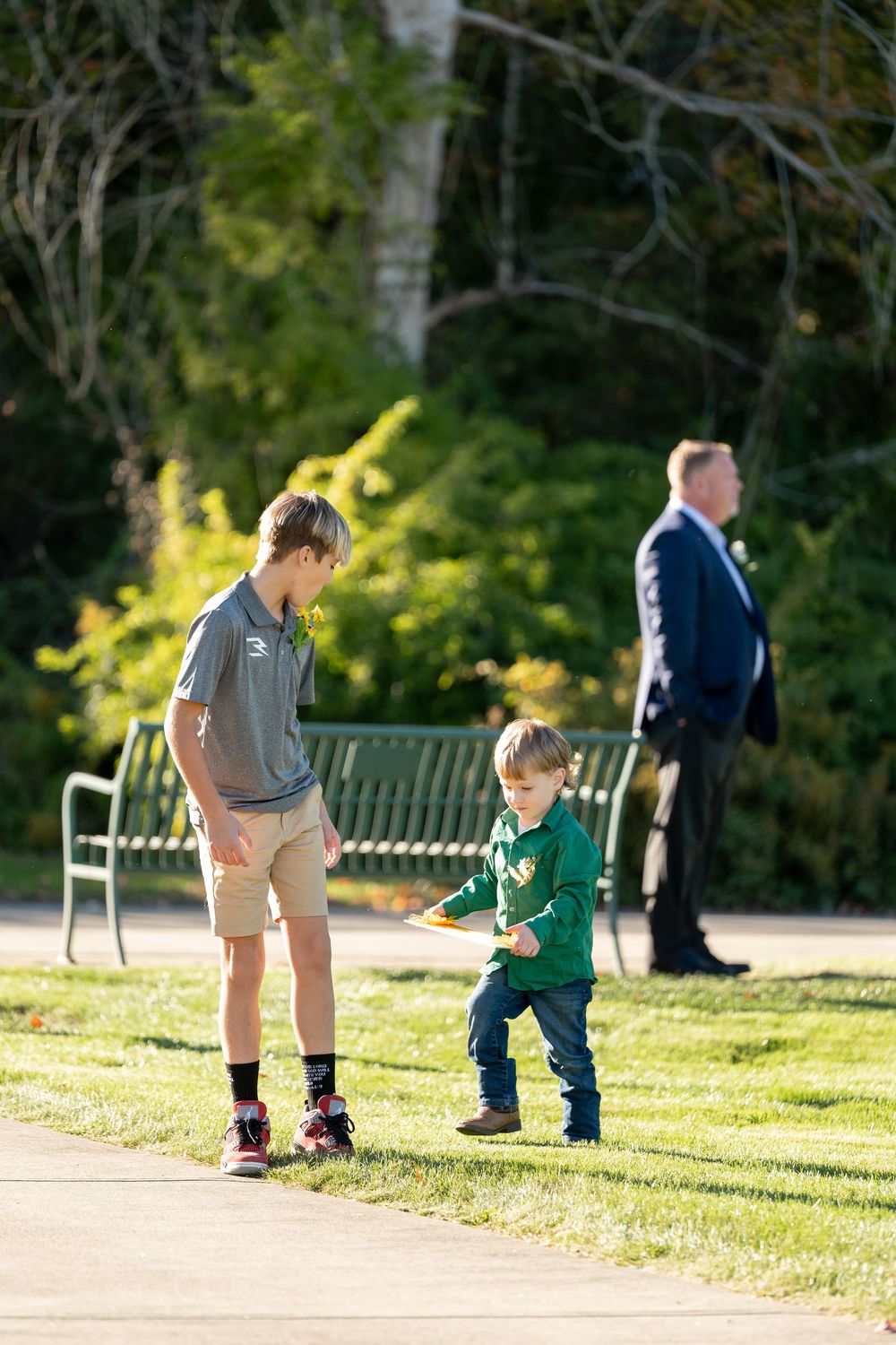 Two boys play on park path near bench while man in suit stands in background at wedding.