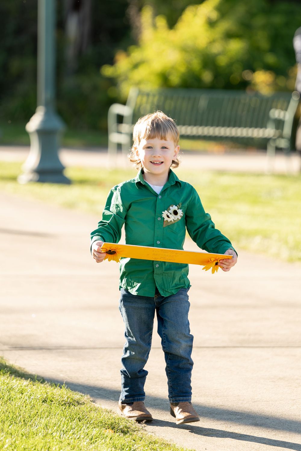 Happy young boy in green shirt and jeans holds yellow sign on sunny park pathway.