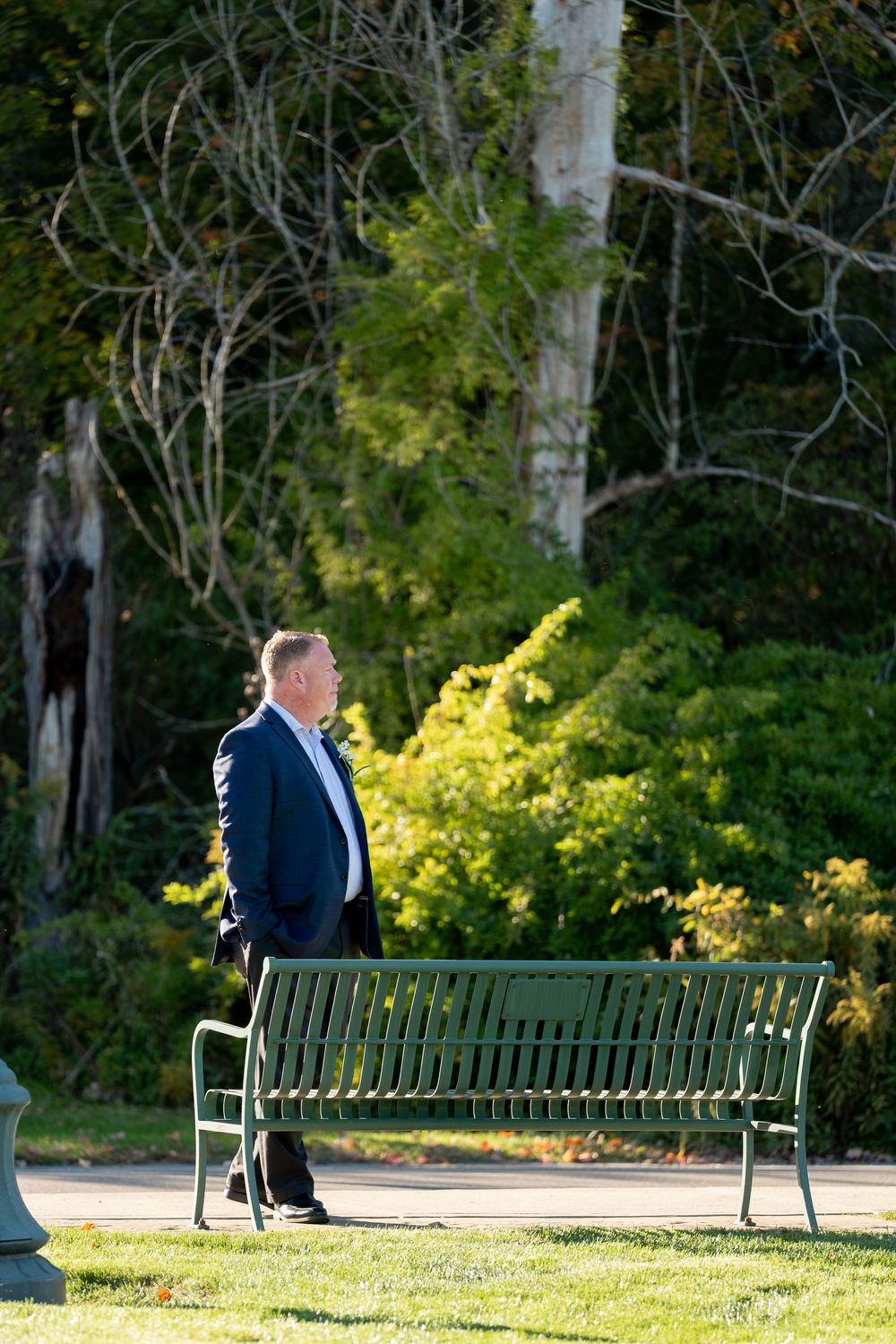Man in blue suit walks past green metal bench in sunlit park setting.