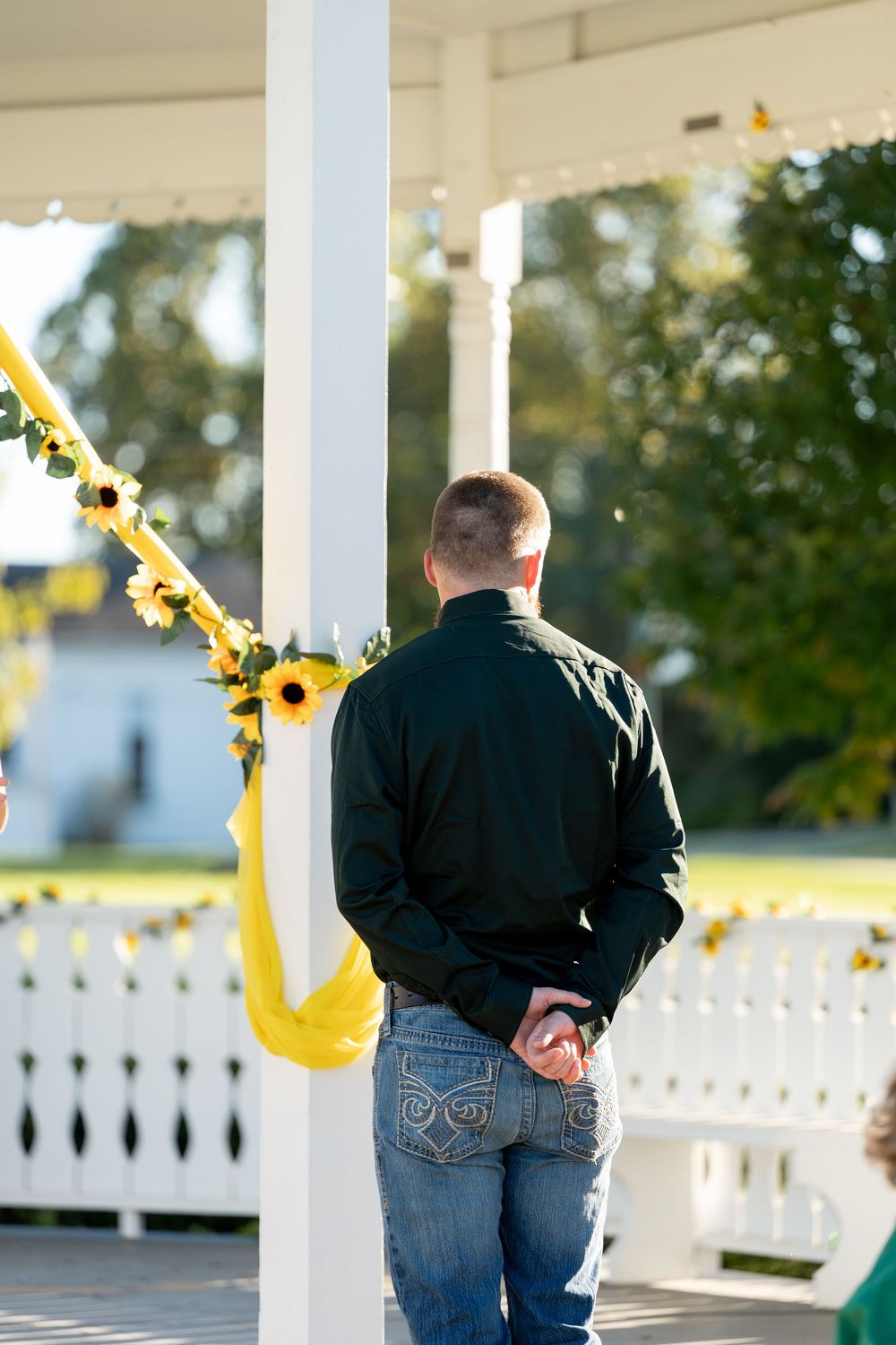 Groom in black shirt and jeans stands at sunflower-decorated white gazebo wedding altar.