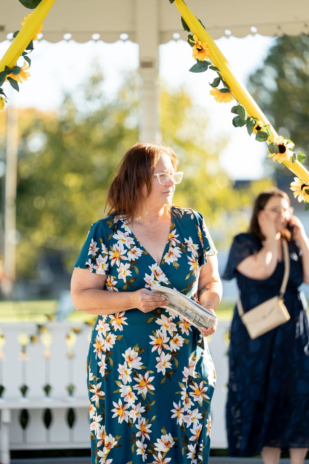 Woman in teal floral dress holds notes while standing under sunflower-decorated gazebo arch.