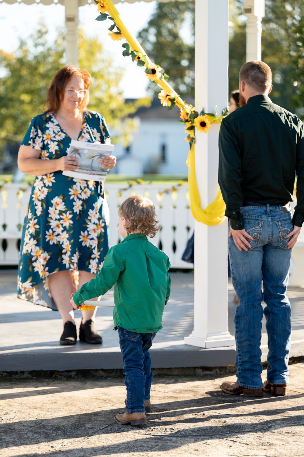 Young boy in green shirt stands beside groom as officiant approaches sunflower-decorated gazebo.