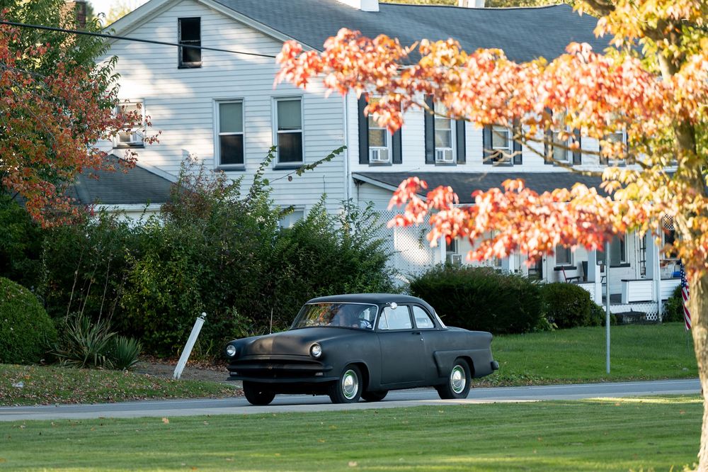 Vintage black classic car driving past a white house with vibrant autumn foliage trees.