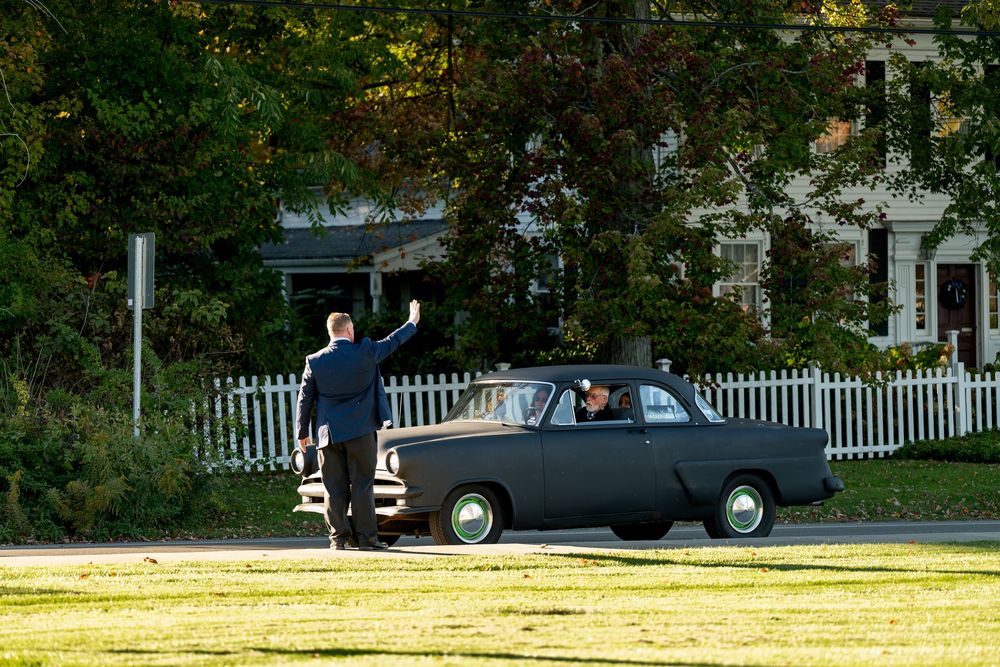 Man waves goodbye to passengers in vintage black car driving across sunny park lawn.