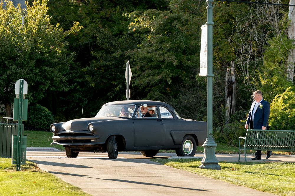 Vintage black car drives through sunny park past lamppost with man watching nearby.