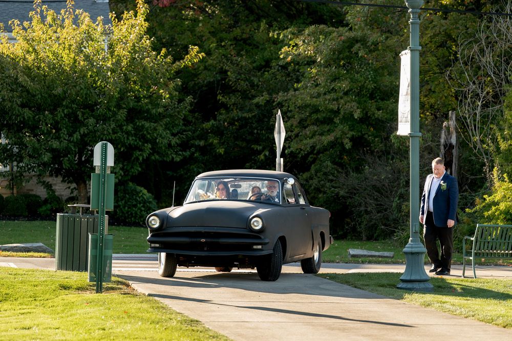 Vintage black classic car arriving at park as man in suit watches on sunny autumn day.