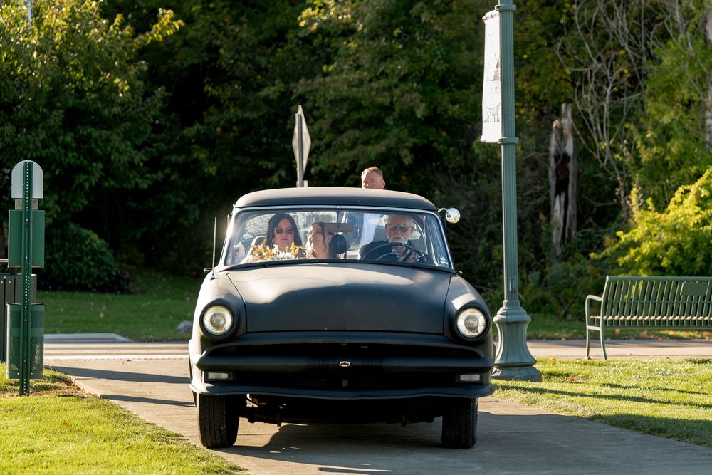 Bride and groom ride in vintage black convertible car through sunny park after wedding.