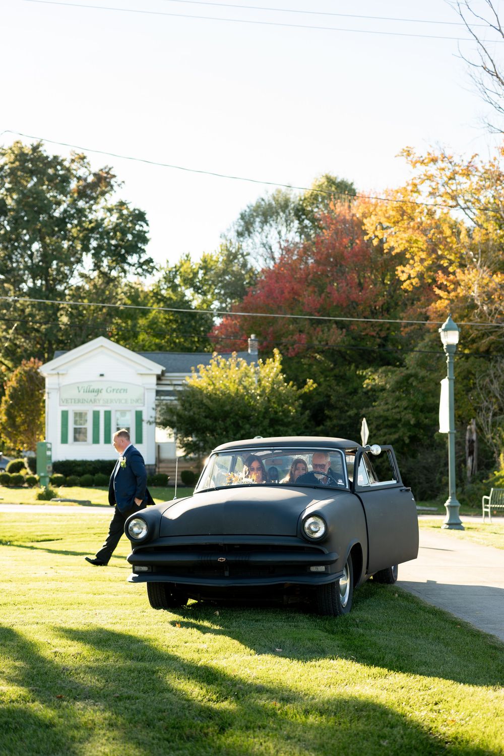 Vintage matte black classic car parked on green lawn outside a white building with autumn trees in background.