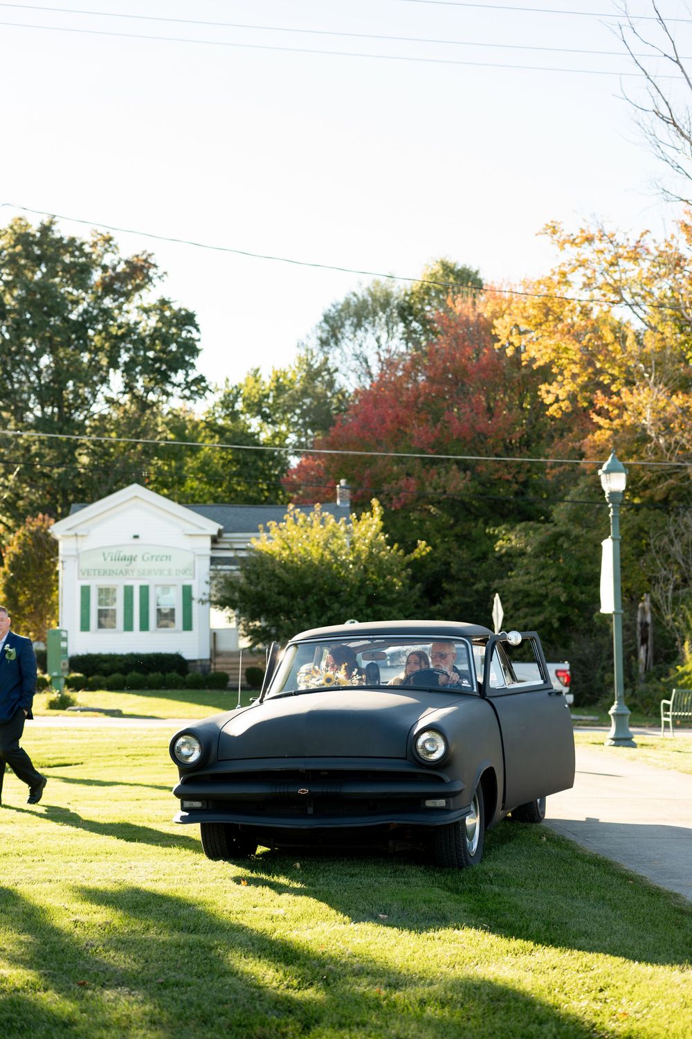 Vintage matte black classic car parked on grass outside a white building with autumn trees in background.