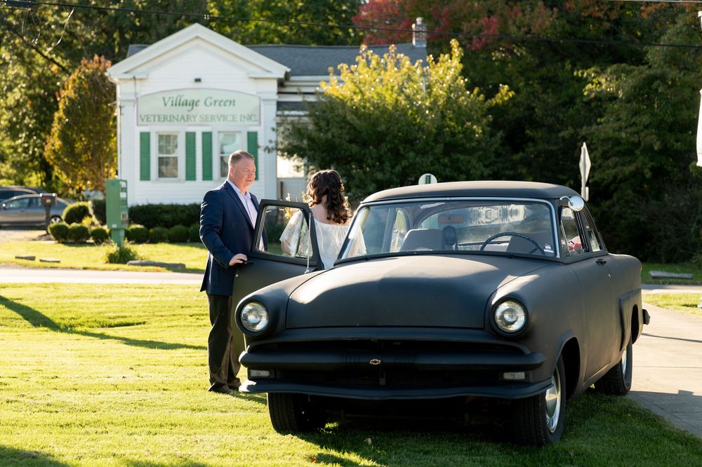 Man in suit stands beside vintage matte black car outside a country club building.