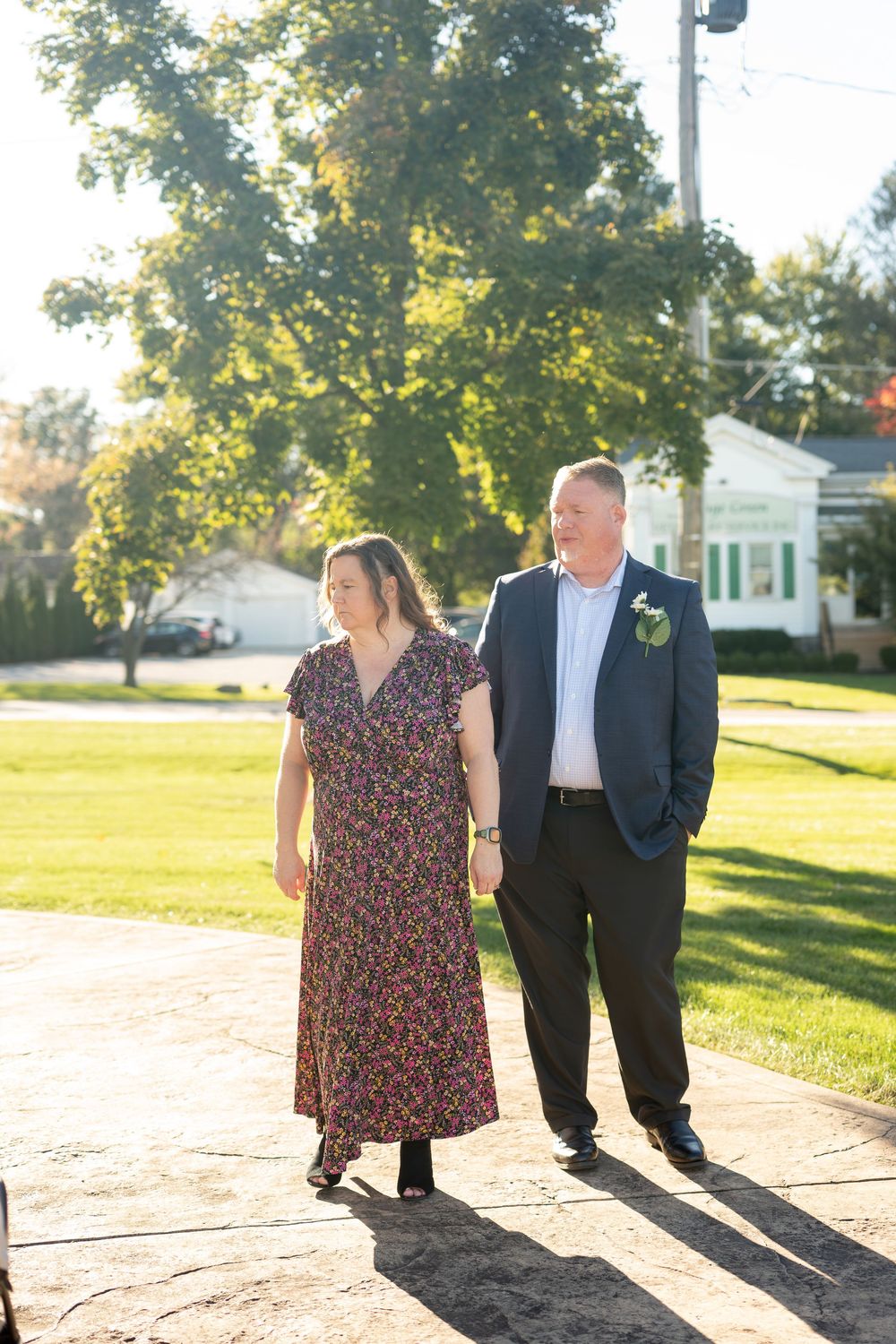 Couple walks hand in hand in warm sunlight outside near a green lawn and white building.