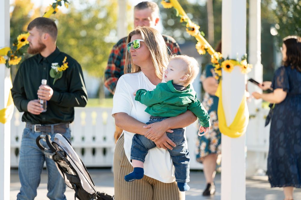 Woman in white top holds baby in green outfit at outdoor sunflower-themed wedding celebration.