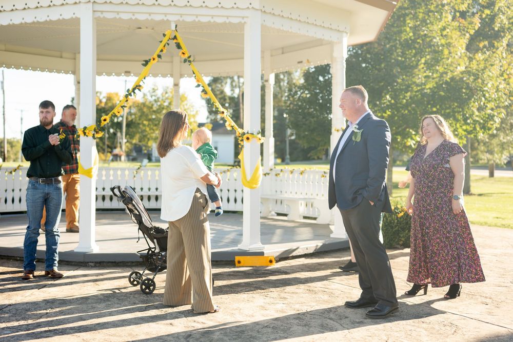 Wedding guests gather near sunflower-decorated gazebo in sunny outdoor park setting.