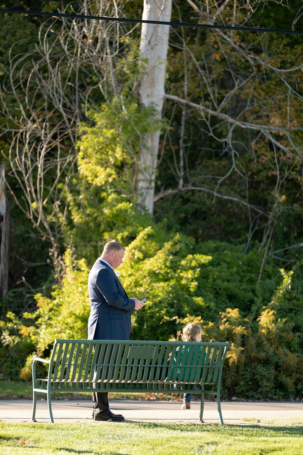 Man in blue suit stands near green park bench surrounded by autumn foliage.