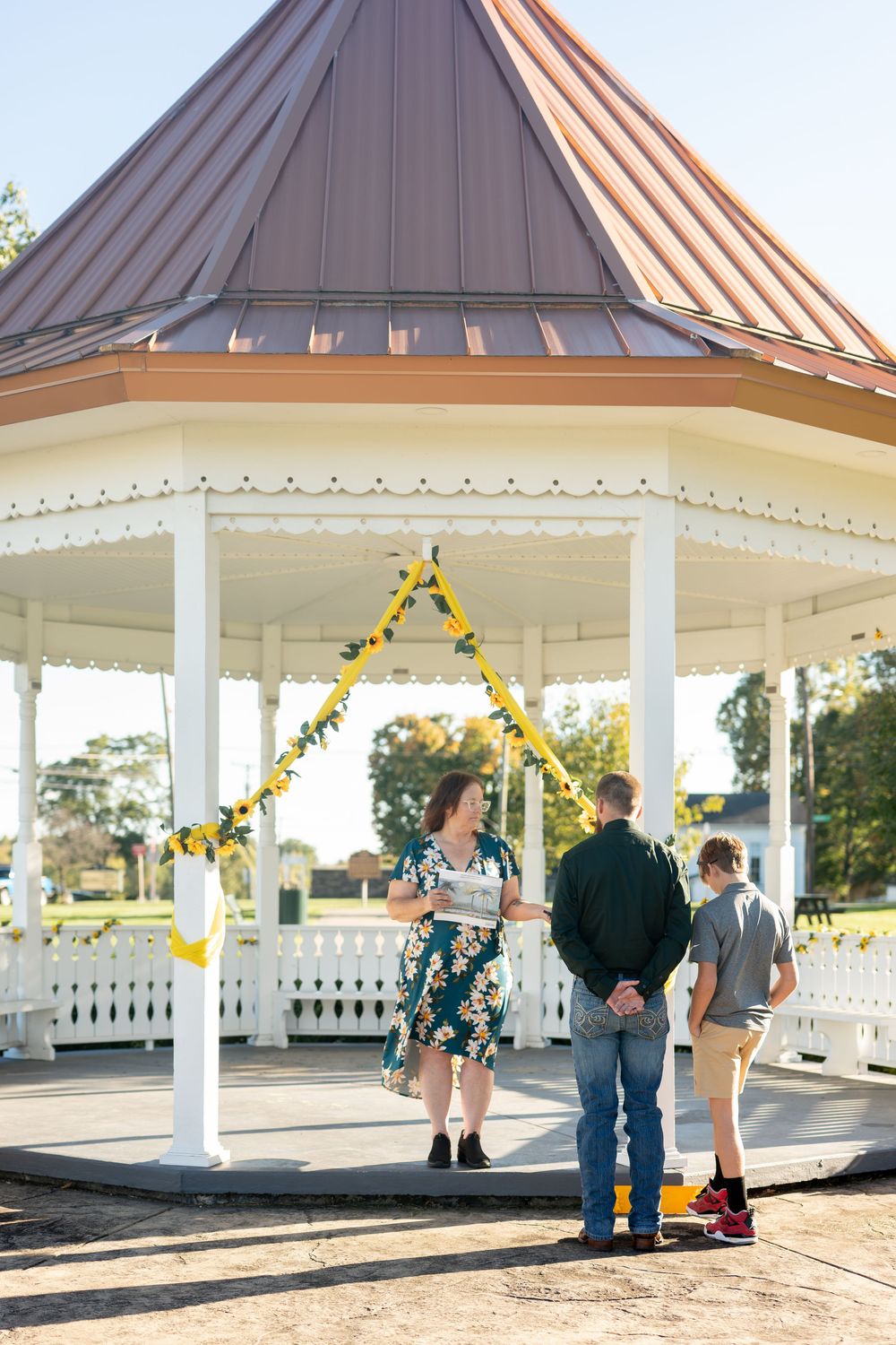 Wedding officiant in floral dress reviews vows with couple at sunflower-decorated white gazebo.