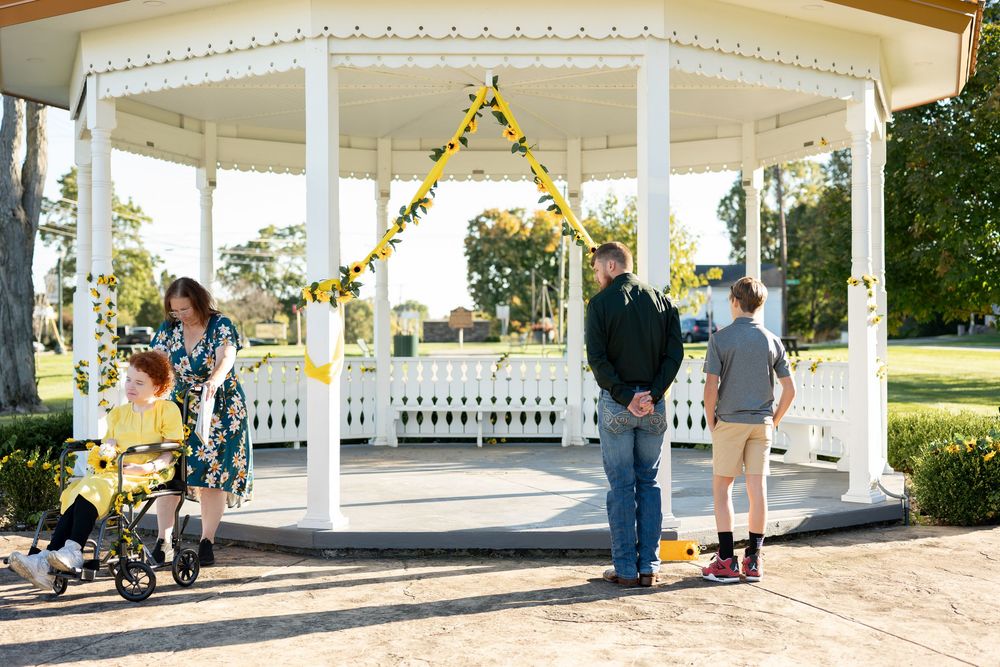 Small wedding gathering at white gazebo decorated with yellow ribbons and sunflower garland arch.