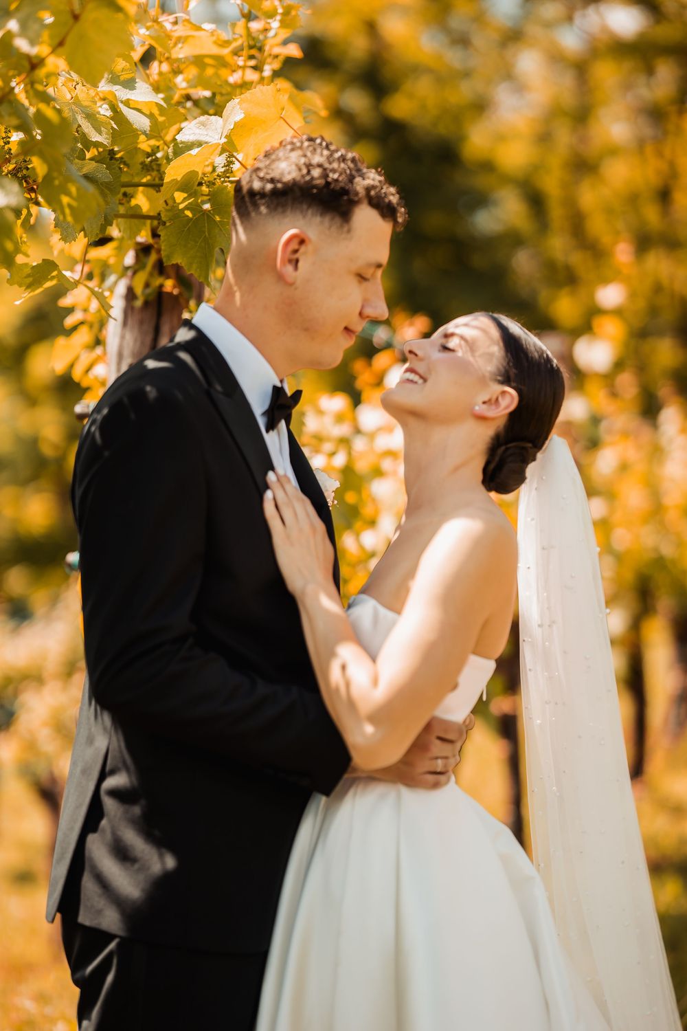 Egy romantikus őszi esküvői fotó, amelyen egy pár ölelkezik a szabadban, aranysárga lombozat között.