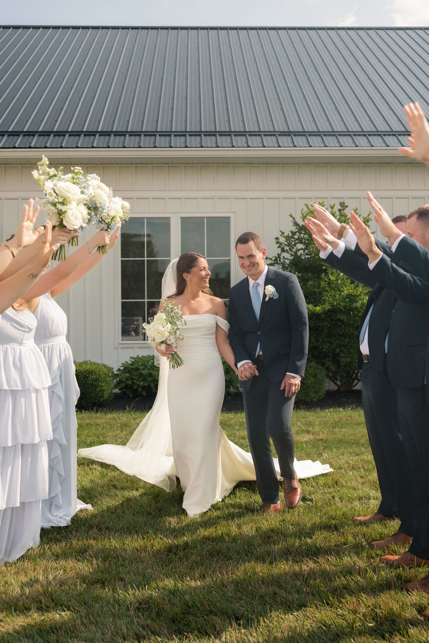 Joyful wedding party in dusty blue attire celebrates with bride and groom outside a white barn venue.