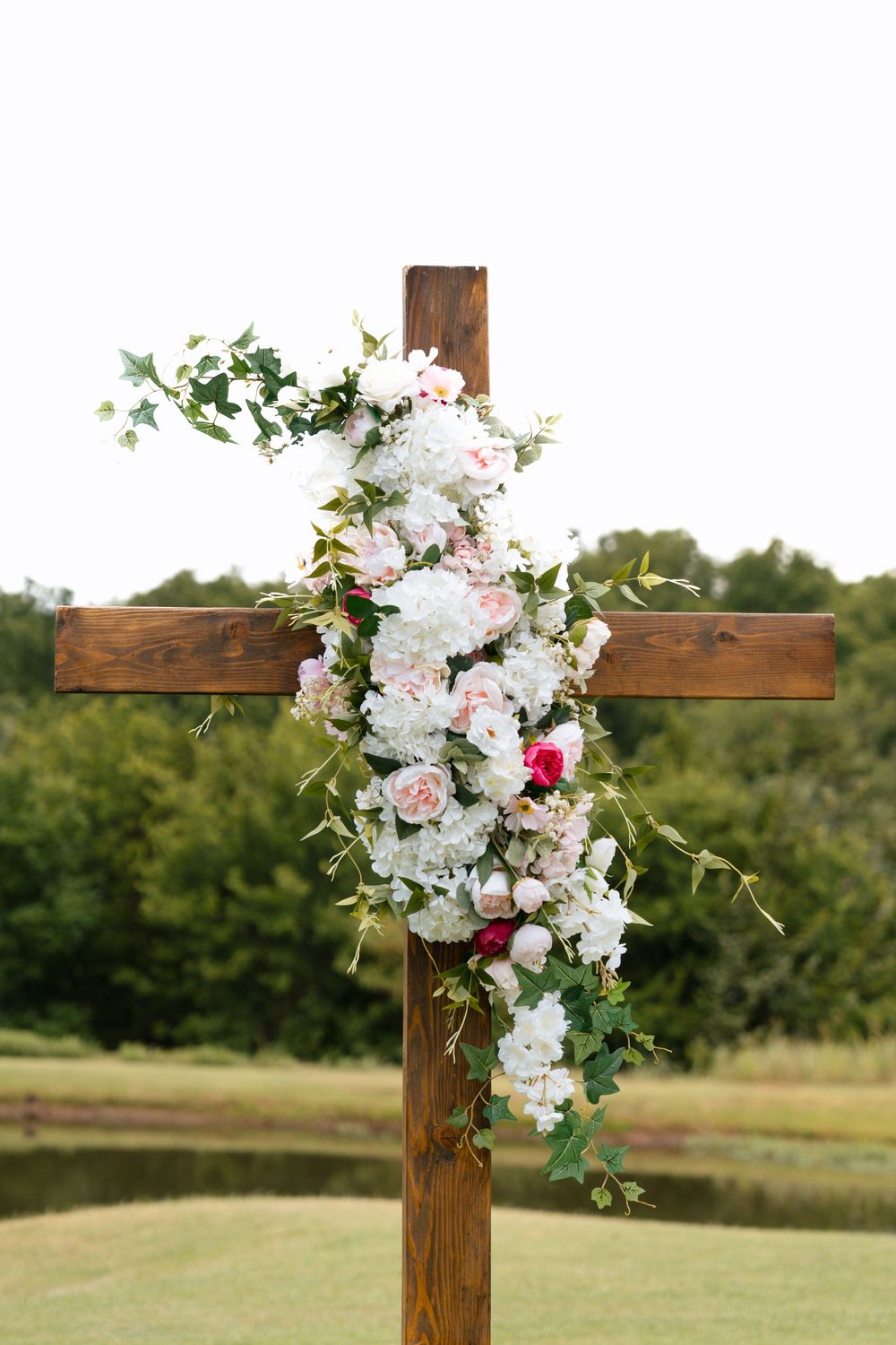 Wooden cross decorated with white and pink floral arrangement and greenery outdoors at a wedding ceremony.