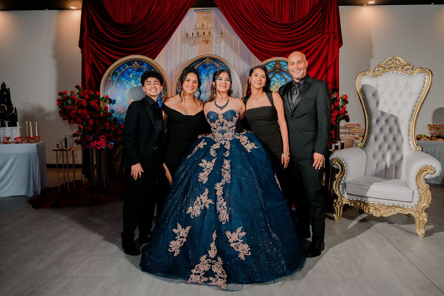 Family picture at a quinceañera, with the honoree in a blue embroidered ball gown beside a white throne chair.