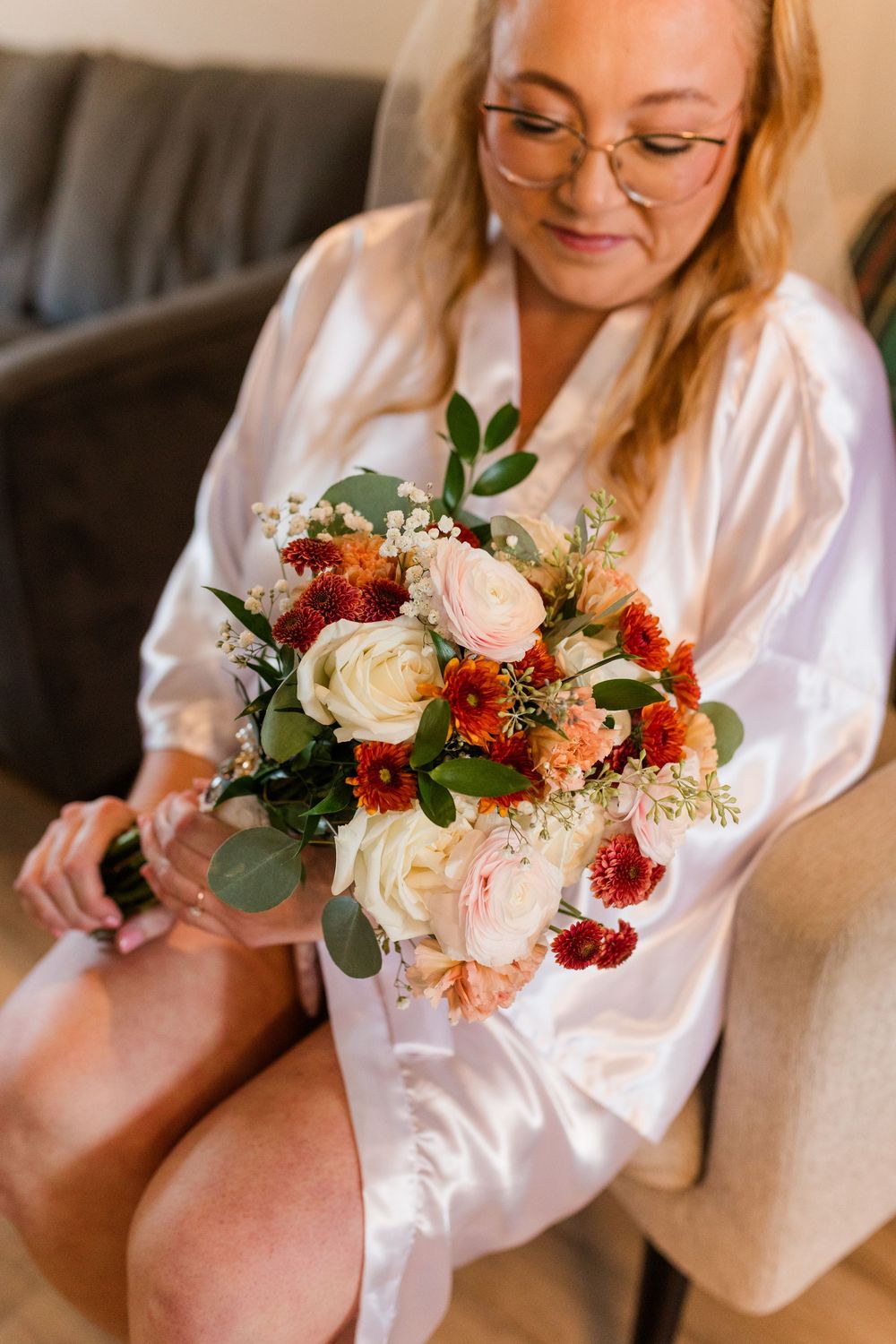 Bride in a white satin robe sits in a chair holding a bouquet of cream roses, orange and red blooms, and eucalyptus greenery.
