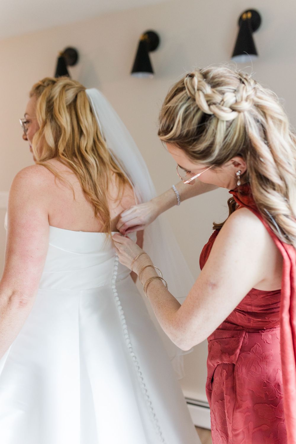 Woman in a red dress with a braided updo fastens the buttons on the back of a bride's white strapless wedding gown and veil.