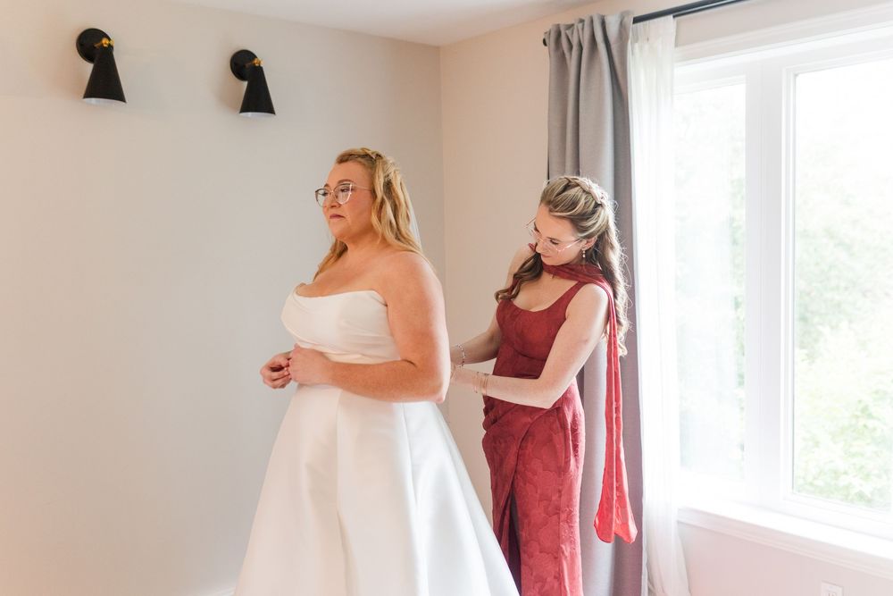 A bridesmaid in a burgundy dress helps a bride in a white strapless wedding gown get ready beside a bright window.