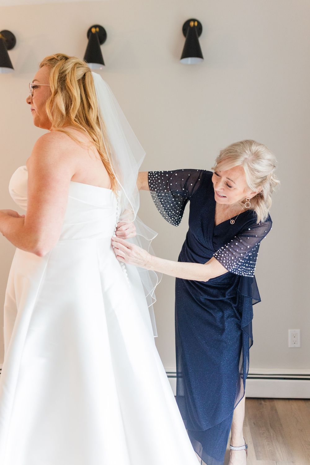 A woman in a navy beaded dress smiles as she helps a bride in a strapless white gown and veil fasten her wedding dress.