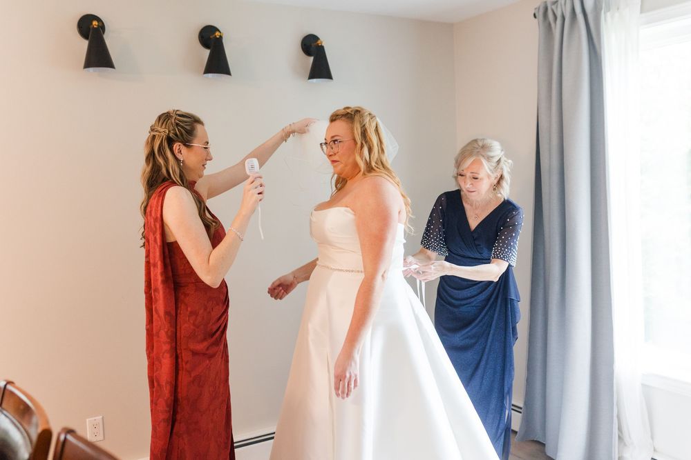 Bride in a strapless white wedding gown getting ready as two women in red and navy dresses help adjust her dress in a bright room.