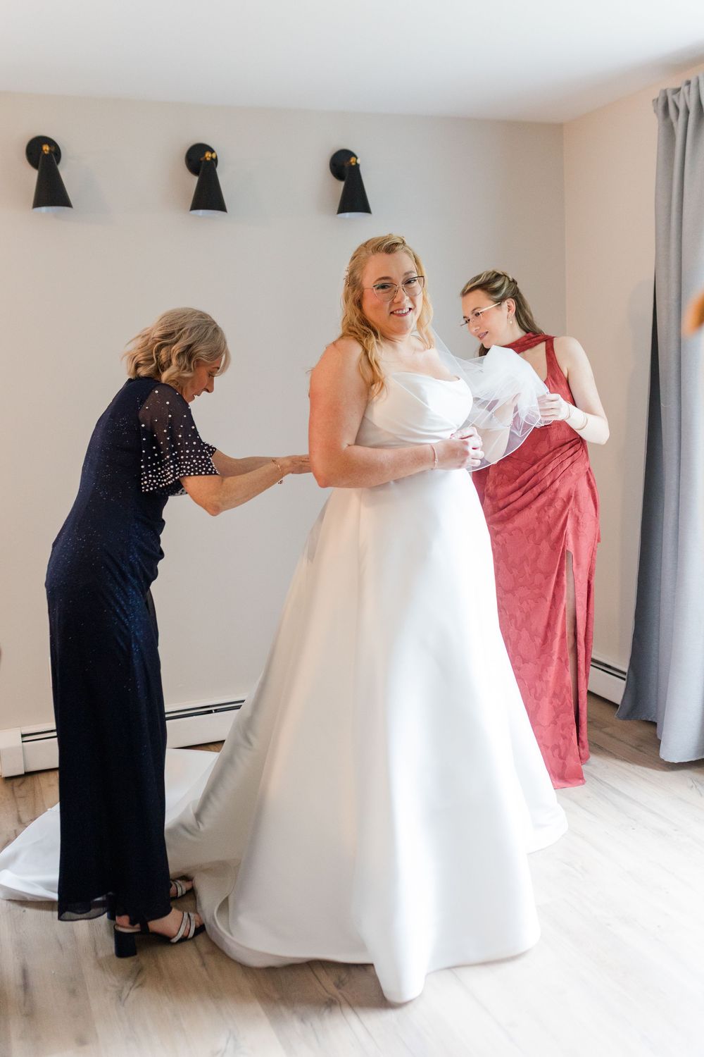 Bride in a white satin wedding gown gets help with final dress adjustments from two women in navy and coral dresses in a bright room.