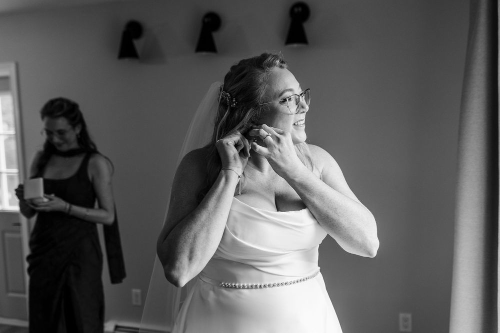 Smiling bride in a white gown and veil adjusts her earring while getting ready, with a woman in black standing in the background.