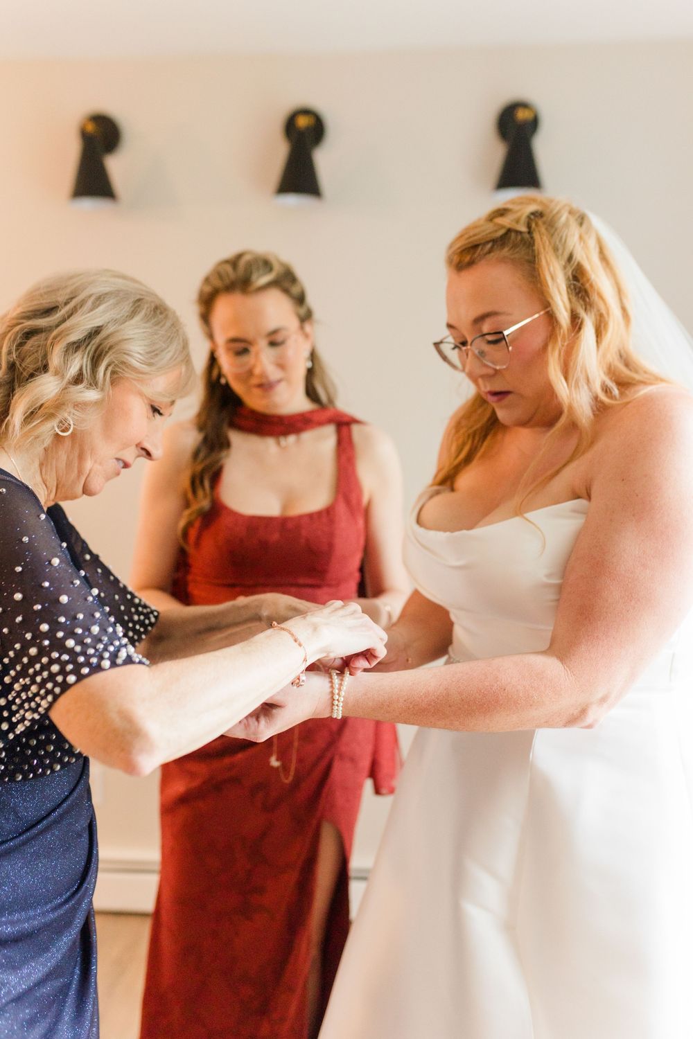A bride in a white strapless gown and veil has her bracelet fastened by an older woman as a bridesmaid in red looks on.
