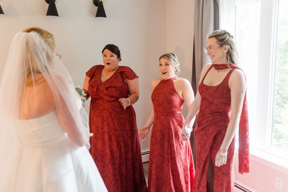 Three bridesmaids in red dresses smile with joy as they see the bride in her white gown and veil on the wedding day.