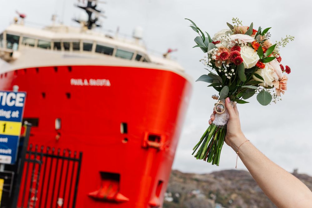 Bride's hand holding a bouquet of red and white flowers in front of a large red cargo ship docked at a harbor.