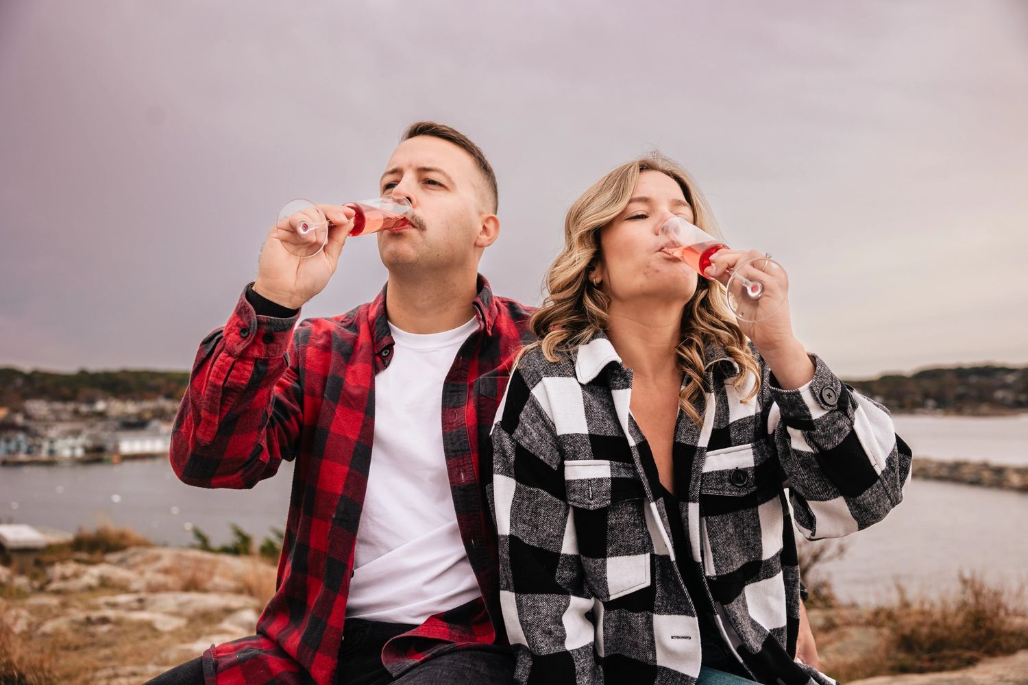 A couple eating popsicles while sitting outdoors overlooking a waterfront, both looking upward. Engagement at Rockport Harbor, Rockport, M