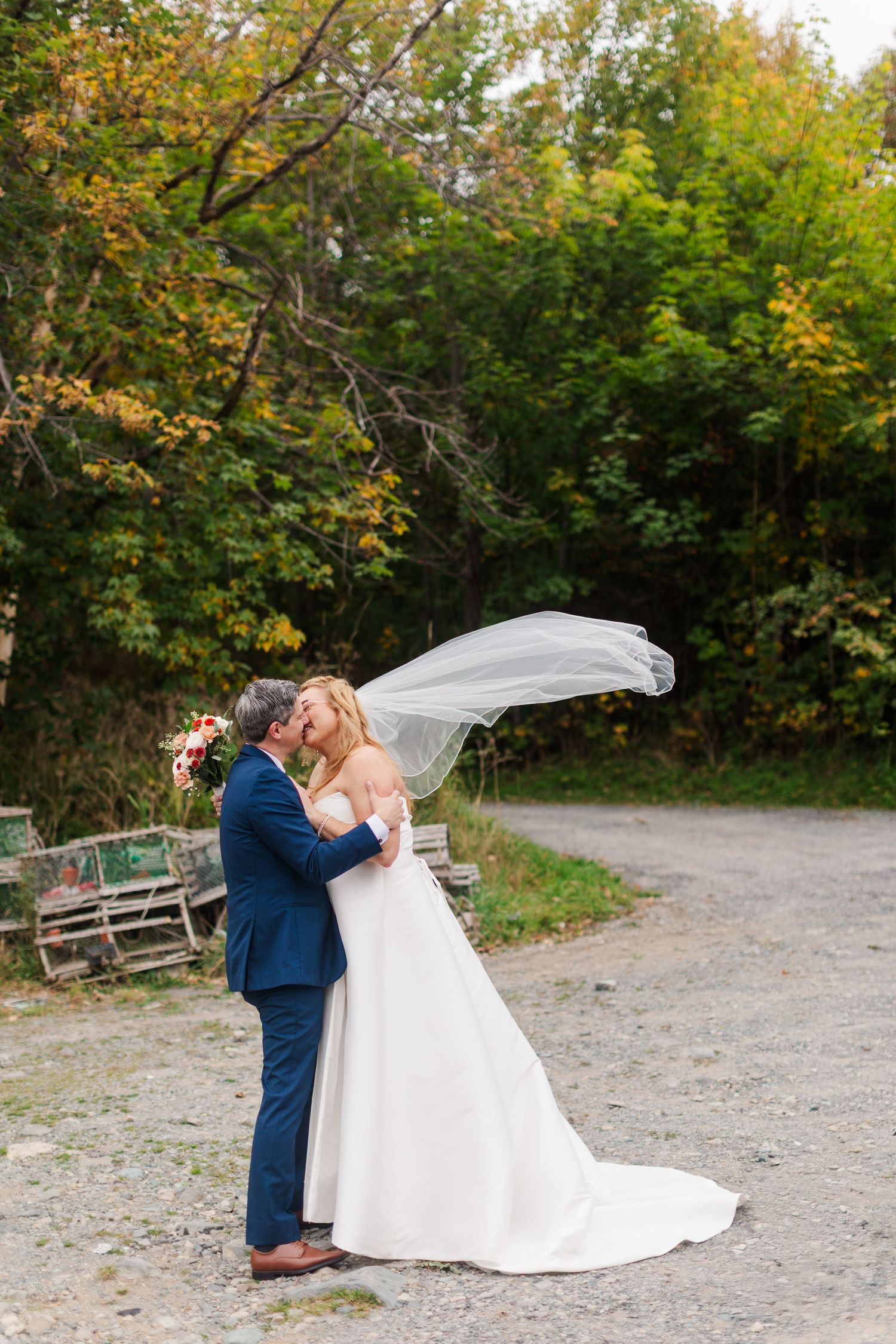 Bride in a white gown and flowing veil kisses groom in a navy suit on a gravel path surrounded by colorful autumn trees.