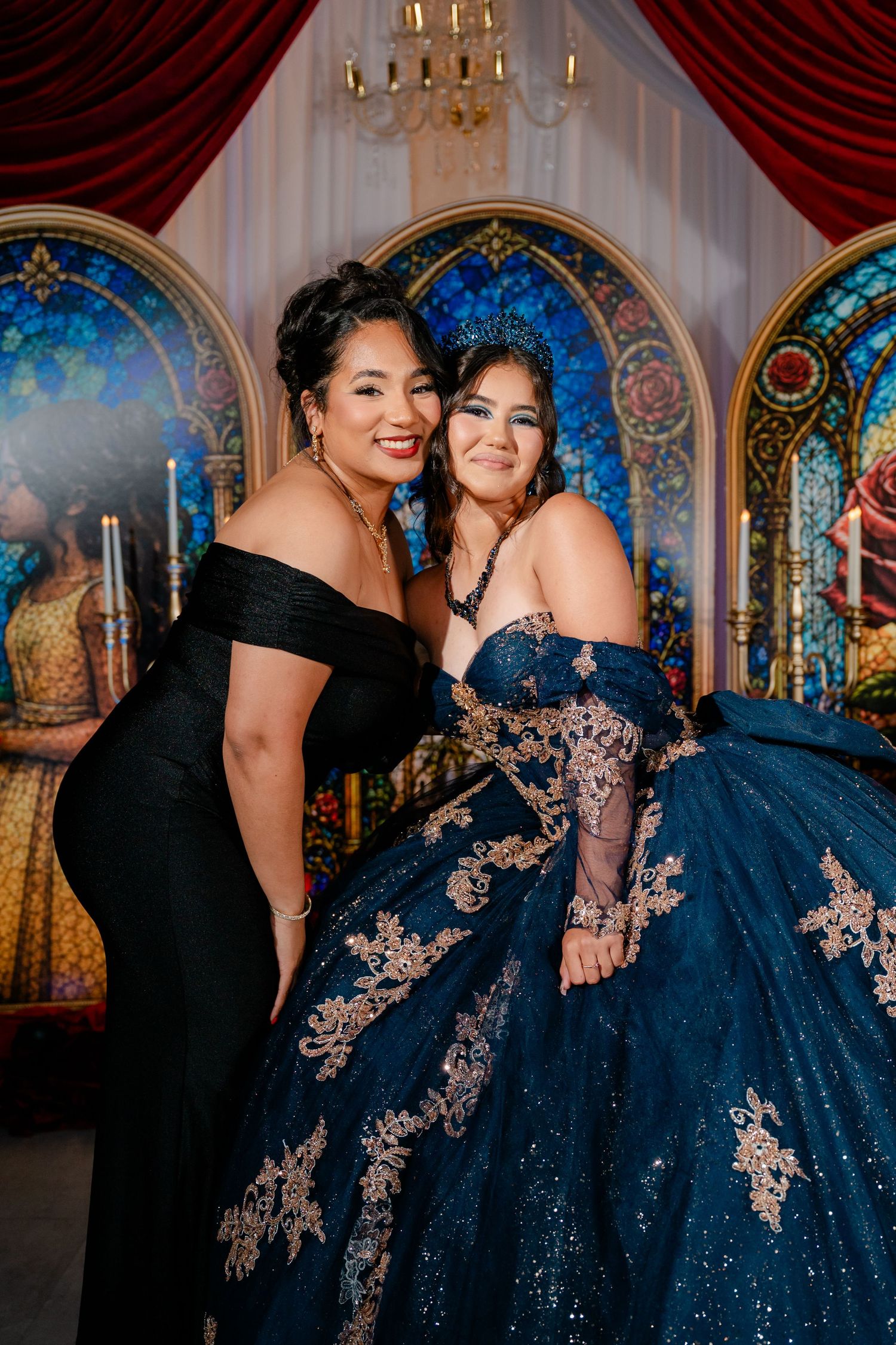 Two smiling women embrace at a quinceañera, one in a black off-shoulder gown and one in a navy gold-embroidered ball gown with a tiara.