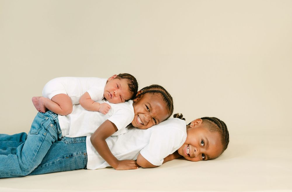 Three children in white shirts stacking together smiling, with a newborn baby on top in a studio portrait.