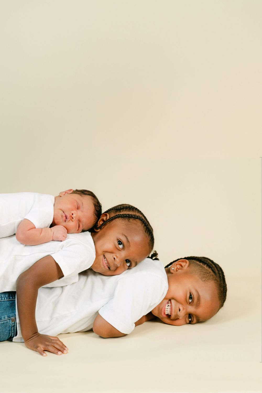 Two smiling children in white shirts pose with a sleeping newborn baby on a beige background.