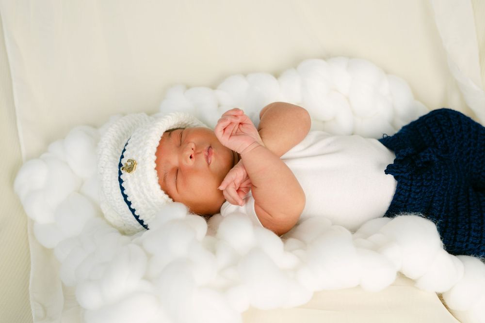 Newborn baby sleeping peacefully in white knit sailor hat and navy outfit, nestled in fluffy white cotton clouds.
