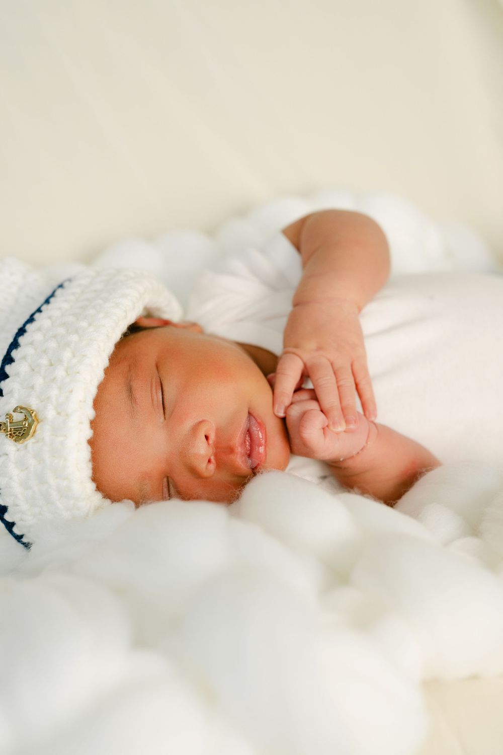 Sleeping newborn baby wearing a white knit sailor hat, lying on soft white blanket with tiny hands visible.