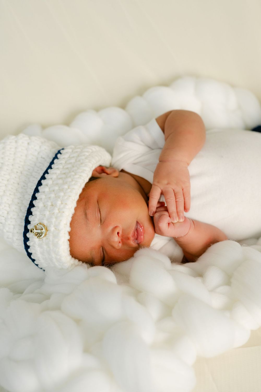 Sleeping newborn baby wearing a white and navy crochet sailor hat, resting on fluffy white cotton backdrop.