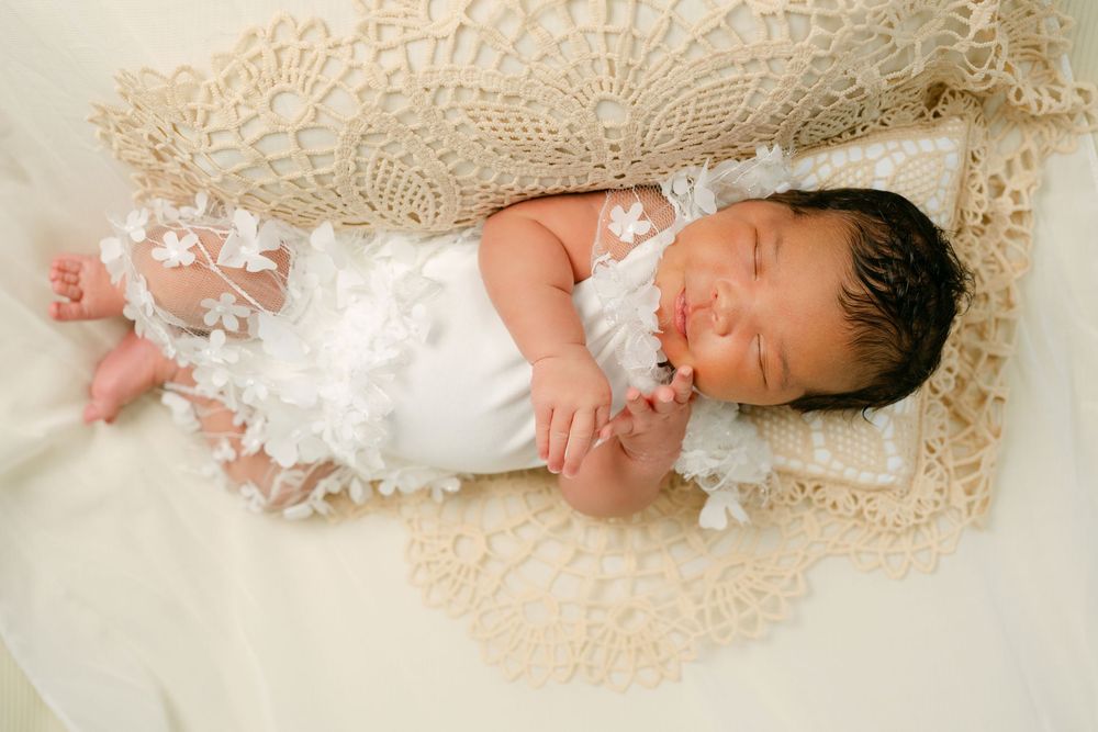 Sleeping newborn baby in white lace outfit on crochet blanket, peaceful infant portrait photography.