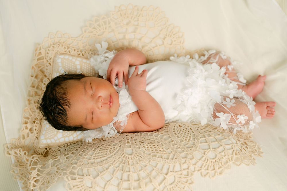 Newborn baby sleeping peacefully in white lace outfit on crochet blanket in a wooden basket.