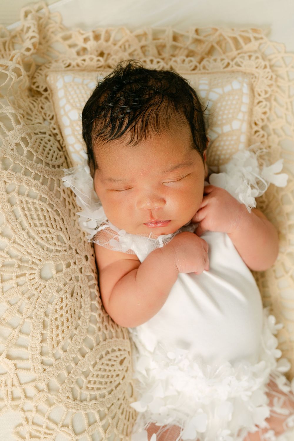 Sleeping newborn baby in white lace outfit resting on cream crochet blanket with delicate pillow.