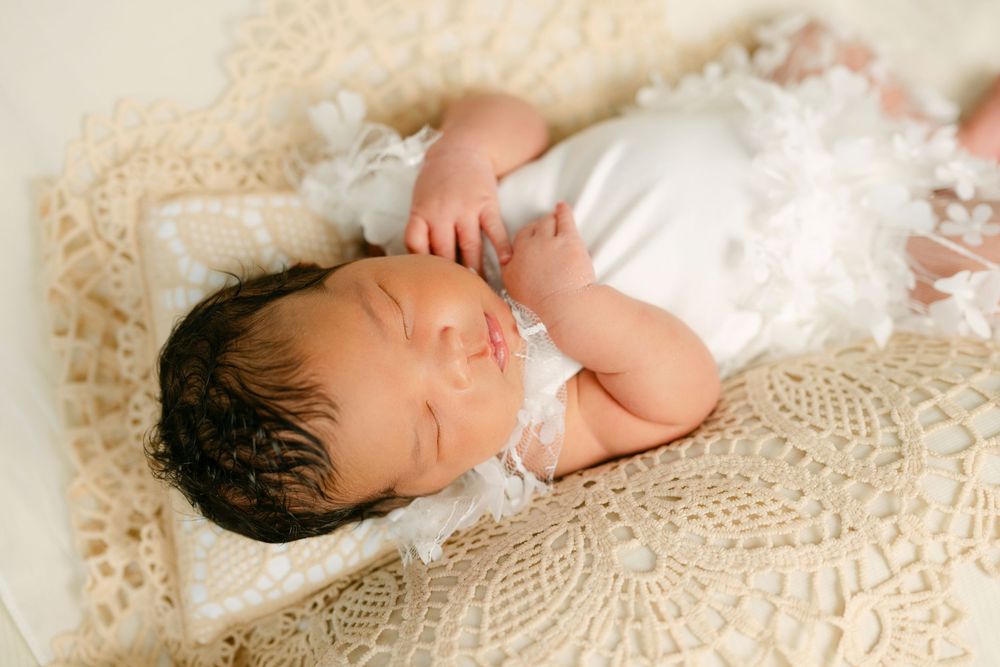 Sleeping newborn baby in white outfit on cream lace blanket, peaceful infant portrait photography.